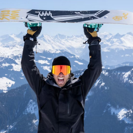 Porträttbild på Henrik, ståendes i skidutrustning lyftandes en snowboard över huvudet